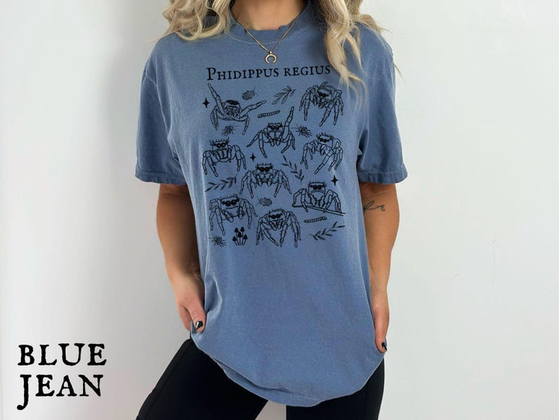Pu&ograve; includere: T-shirt blu jeans con illustrazione nera di ragni saltatori e la scritta "PHIDIPPUS REGIUS". La maglietta ha una vestibilit&agrave; ampia e maniche corte, adatta per un abbigliamento casual.