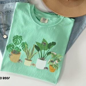 May include: A light green t-shirt with a graphic of potted plants and small spiders. The text "Island Reef" is printed at the bottom of the shirt.