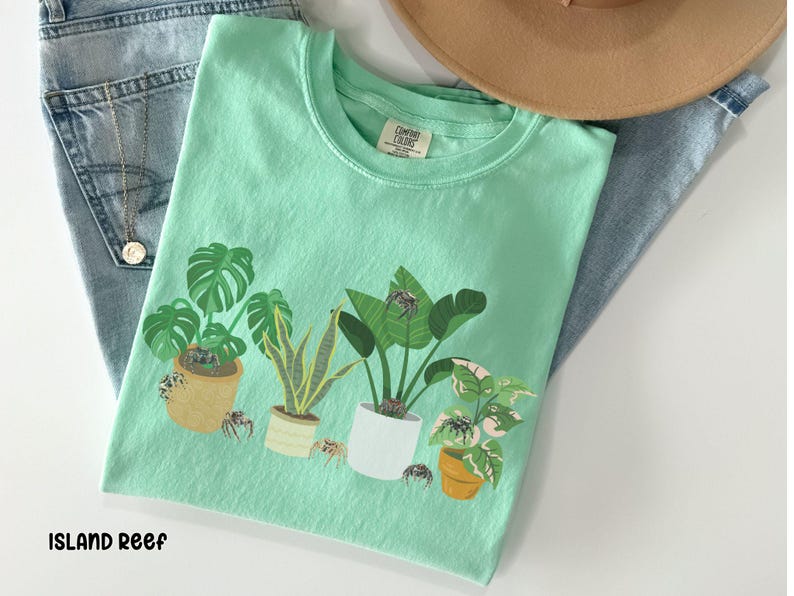 May include: A light green t-shirt with a graphic of potted plants and small spiders. The text "Island Reef" is printed at the bottom of the shirt.
