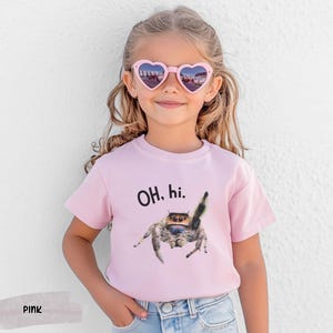 May include: A pink t-shirt with a black and white jumping spider graphic and the text "OH, hi." printed on the front. The shirt is being worn by a child wearing pink heart-shaped sunglasses.