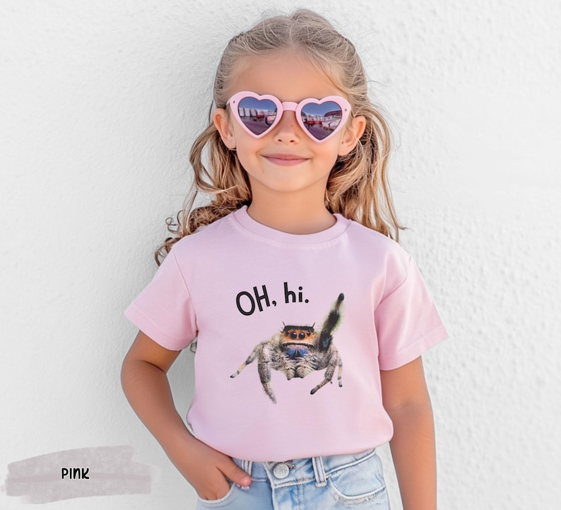 May include: A pink t-shirt with a black and white jumping spider graphic and the text "OH, hi." printed on the front. The shirt is being worn by a child wearing pink heart-shaped sunglasses.