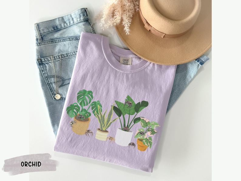 May include: A lavender t-shirt with a graphic of potted plants and spiders. The text "ORCHID" is printed on the bottom left corner of the image.