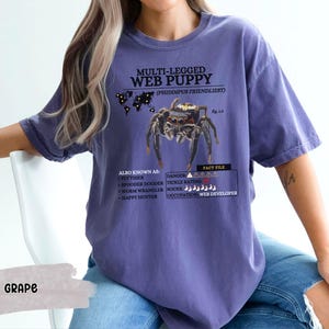 May include: Purple t-shirt with a graphic of a spider and the text "MULTI-LEGGED WEB PUPPY (PHIDDIPUS FRIENDLIEST)". The text also includes "ALSO KNOWN AS: FLY TIMER, SPOODER DOODER, WORM WRANGLER, HAPPY HUNTER" and "FACT FILE: DANGER, TICKLE RATING, OCCUPATION: WEB DEVELOPER".