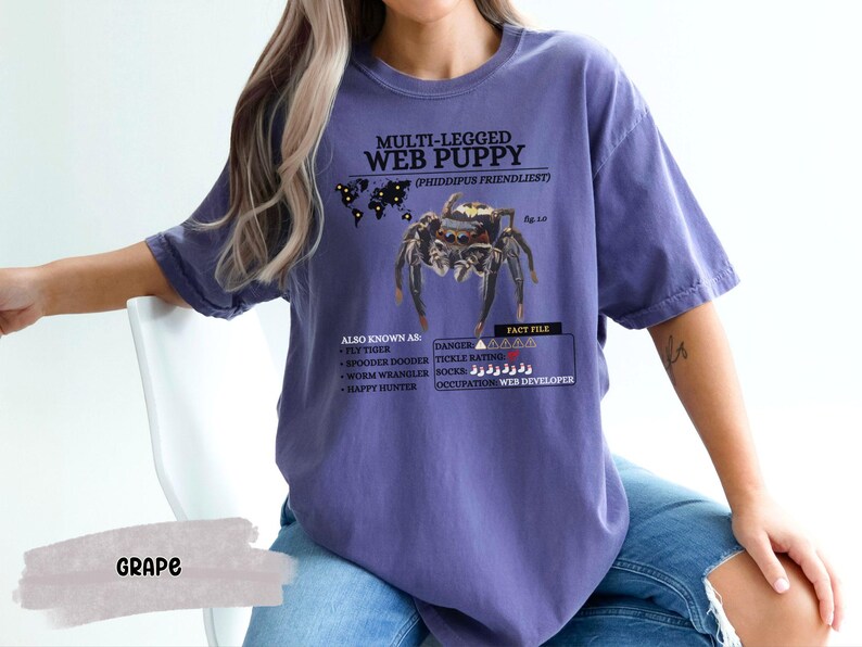 May include: Purple t-shirt with a graphic of a spider and the text "MULTI-LEGGED WEB PUPPY (PHIDDIPUS FRIENDLIEST)". The text also includes "ALSO KNOWN AS: FLY TIMER, SPOODER DOODER, WORM WRANGLER, HAPPY HUNTER" and "FACT FILE: DANGER, TICKLE RATING, OCCUPATION: WEB DEVELOPER".
