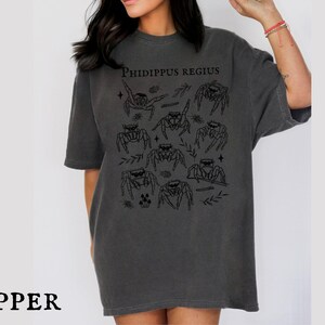 Pu&ograve; includere: T-shirt grigio scuro con una grafica di ragni saltatori neri e la scritta "Phidippus Regius". La maglietta ha una vestibilit&agrave; ampia e maniche corte. La parola "PEPPER" &egrave; in basso.