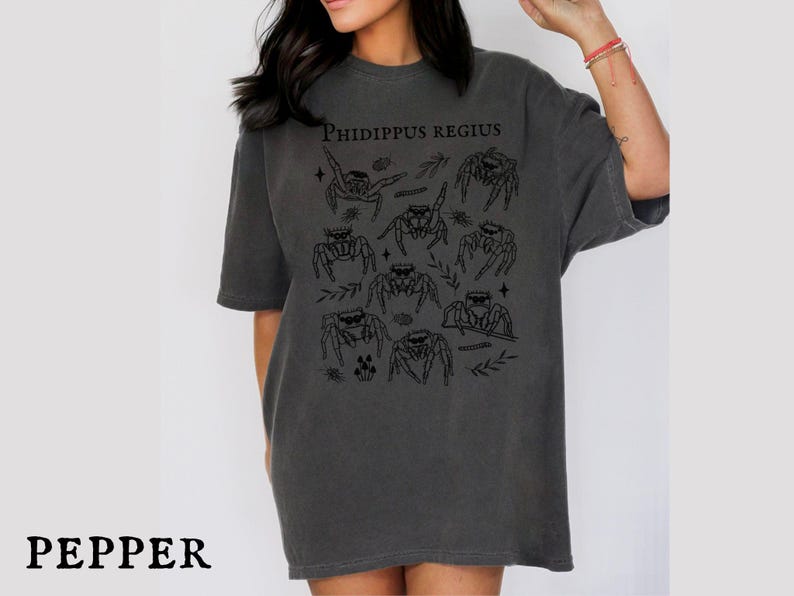 Pu&ograve; includere: T-shirt grigio scuro con una grafica di ragni saltatori neri e la scritta "Phidippus Regius". La maglietta ha una vestibilit&agrave; ampia e maniche corte. La parola "PEPPER" &egrave; in basso.