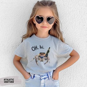 May include: A young person wearing a grey t-shirt with the text "OH, hi." and a jumping spider graphic. The person is wearing heart-shaped sunglasses and blue jeans.
