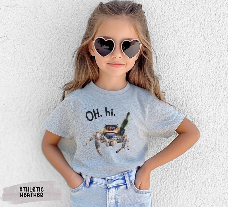 May include: A young person wearing a grey t-shirt with the text "OH, hi." and a jumping spider graphic. The person is wearing heart-shaped sunglasses and blue jeans.
