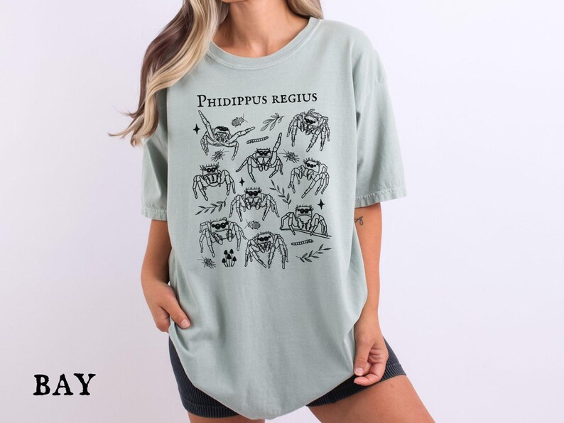 Pu&ograve; includere: T-shirt verde salvia con la scritta "Phidippus Regius" e un disegno in bianco e nero di diversi ragni saltatori. I ragni sono raffigurati in varie pose. La maglietta ha una vestibilit&agrave; ampia.