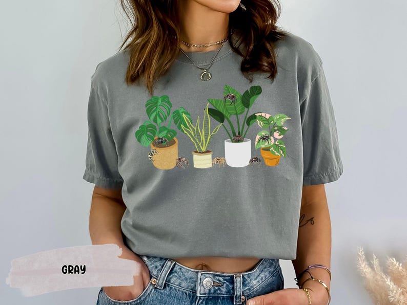 May include: Gray t-shirt with a graphic of potted plants and spiders. The shirt has a crew neck and short sleeves.