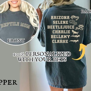 May include: A dark gray t-shirt with the text "REPTILE MOM" on the front. The back of the shirt features the names "ARIZONA, SELENE, BEETLEJUICE, CHARLIE, BELLAMY, CLARKE" with reptile illustrations. The shirt also has the text "100% PERSONALIZED WITH YOUR PETS".