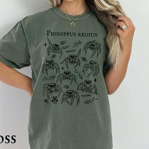 Pu&ograve; includere: T-shirt verde muschio con grafica nera di ragni saltatori e la scritta "Phidippus Regius". La maglietta ha girocollo e maniche corte. La parola "MOSS" &egrave; stampata in basso.