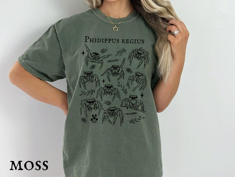 Pu&ograve; includere: T-shirt verde muschio con grafica nera di ragni saltatori e la scritta "Phidippus Regius". La maglietta ha girocollo e maniche corte. La parola "MOSS" &egrave; stampata in basso.