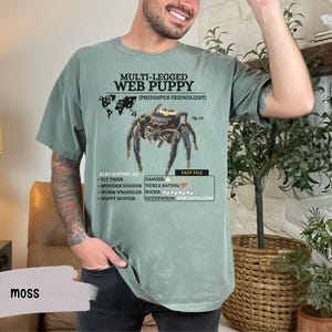 May include: A green t-shirt with a graphic of a jumping spider and the text "MULTI-LEGGED WEB PUPPY (PHIDDIPUS FRIENDLIEST)". The text below the spider graphic reads "ALSO KNOWN AS: FLY TIGER, SPOODER DOODER, WORM WRANGLER, HAPPY HUNTER" and "DANGER: TICKLE RATING: 5/5, OCCUPATION: WEB DEVELOPER".