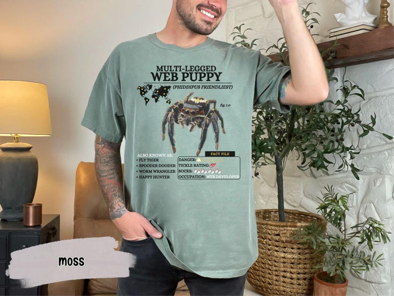 May include: A green t-shirt with a graphic of a jumping spider and the text "MULTI-LEGGED WEB PUPPY (PHIDDIPUS FRIENDLIEST)". The text below the spider graphic reads "ALSO KNOWN AS: FLY TIGER, SPOODER DOODER, WORM WRANGLER, HAPPY HUNTER" and "DANGER: TICKLE RATING: 5/5, OCCUPATION: WEB DEVELOPER".