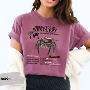 May include: A purple t-shirt with a graphic of a spider and the text "Multi-legged Web Puppy (Phiddipus Friendliest)". The graphic also includes a map of the world with pins indicating the spider's location. The text "Also Known As: Fly Tiger, Spooder Dooder, Worm Wrangler, Happy Hunter" is listed below the spider graphic. The text "Fact File: Danger, Tickle Rating, Rocks, Occupation: Web Developer" is listed below the spider graphic.