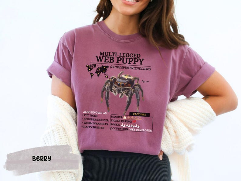 May include: A purple t-shirt with a graphic of a spider and the text "Multi-legged Web Puppy (Phiddipus Friendliest)". The graphic also includes a map of the world with pins indicating the spider's location. The text "Also Known As: Fly Tiger, Spooder Dooder, Worm Wrangler, Happy Hunter" is listed below the spider graphic. The text "Fact File: Danger, Tickle Rating, Rocks, Occupation: Web Developer" is listed below the spider graphic.