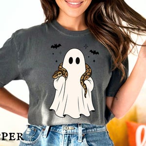 May include: A charcoal gray short-sleeve t-shirt featuring a graphic of a white ghost draped in a sheet with a brown and black patterned snake wrapped around its shoulders, accompanied by two small black bats and four-pointed star accents.