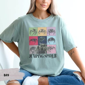 Op de afbeelding: Een lichtblauw t-shirt met een afbeelding van springende spinnen in een kleurrijk rasterpatroon. De tekst "I'm a Jumping Spider" is onder de afbeelding gedrukt.