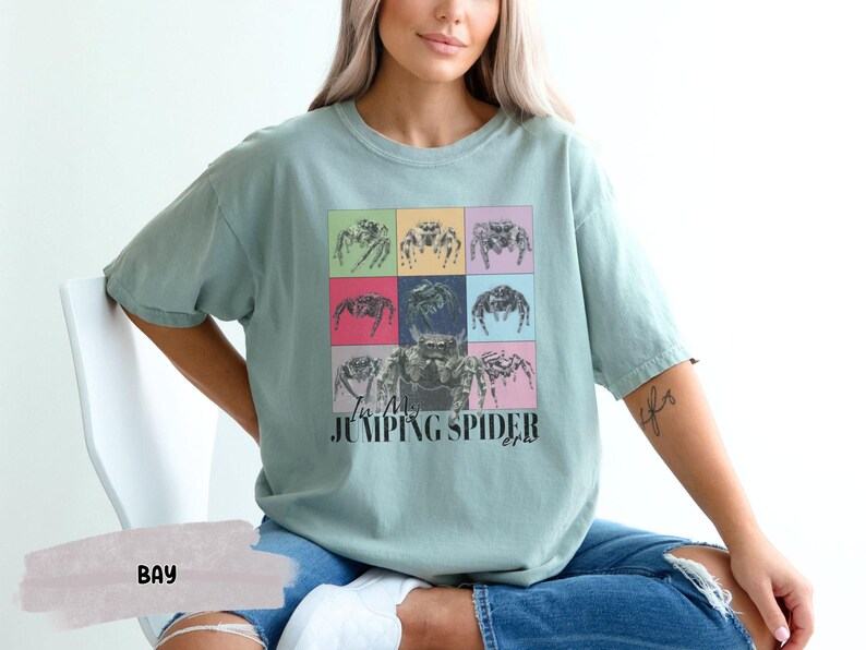 Op de afbeelding: Een lichtblauw t-shirt met een afbeelding van springende spinnen in een kleurrijk rasterpatroon. De tekst "I'm a Jumping Spider" is onder de afbeelding gedrukt.