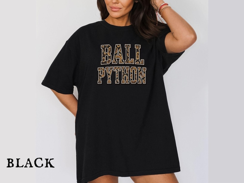 Puede incluir: Una camiseta negra de manga corta con el texto BALL PYTHON impreso en una fuente de bloque. Las letras tienen un patr&oacute;n de leopardo marr&oacute;n y negro. La camiseta tiene cuello redondo y un corte holgado, con la palabra BLACK impresa debajo.