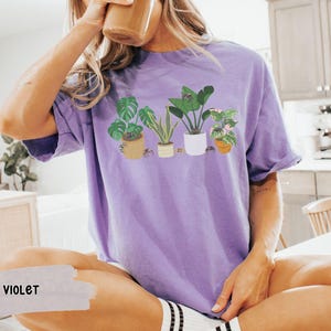 May include: A purple t-shirt with a graphic of potted plants. The shirt has a crew neck and short sleeves.