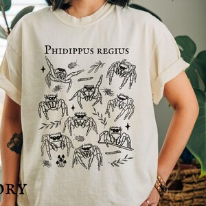 Pu&ograve; includere: T-shirt color avorio con grafica nera di ragni saltatori, foglie e la scritta "Phidippus Regius". Il design include varie illustrazioni di ragni ed elementi botanici. La parola "IVORY" &egrave; stampata in basso.