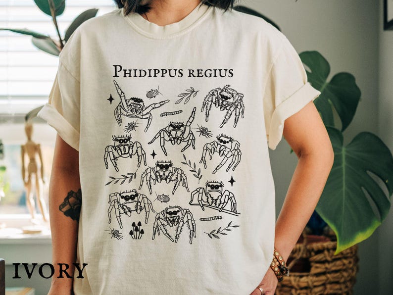 Pu&ograve; includere: T-shirt color avorio con grafica nera di ragni saltatori, foglie e la scritta "Phidippus Regius". Il design include varie illustrazioni di ragni ed elementi botanici. La parola "IVORY" &egrave; stampata in basso.