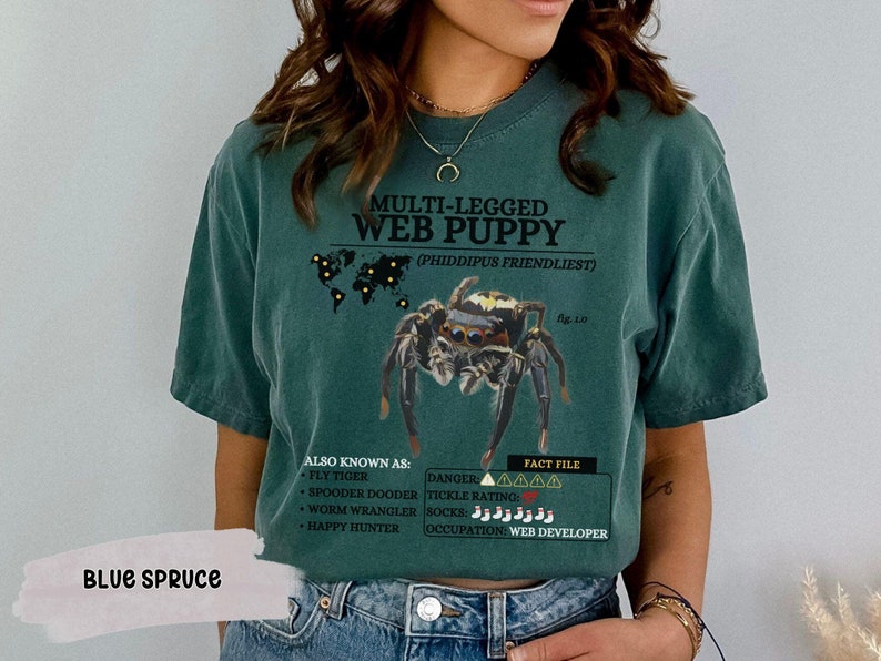 May include: A green t-shirt with a graphic of a jumping spider and the text "MULTI-LEGGED WEB PUPPY (PHIDDIPUS FRIENDLIEST)". The text "ALSO KNOWN AS: FLY TIGER, SPOODER DOODER, WORM WRANGLER, HAPPY HUNTER" and "FACT FILE: DANGER AAAAA, TICKLE RATING: SOCKS:  OCCUPATION: WEB DEVELOPER" is also included on the shirt. The shirt has the text "BLUE SPRUCE" on the bottom left corner.