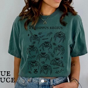Pu&ograve; includere: T-shirt blu abete con un'illustrazione nera di ragni saltatori e la scritta "Phidippus Regius". Il design include elementi botanici. La maglietta ha una vestibilit&agrave; ampia.