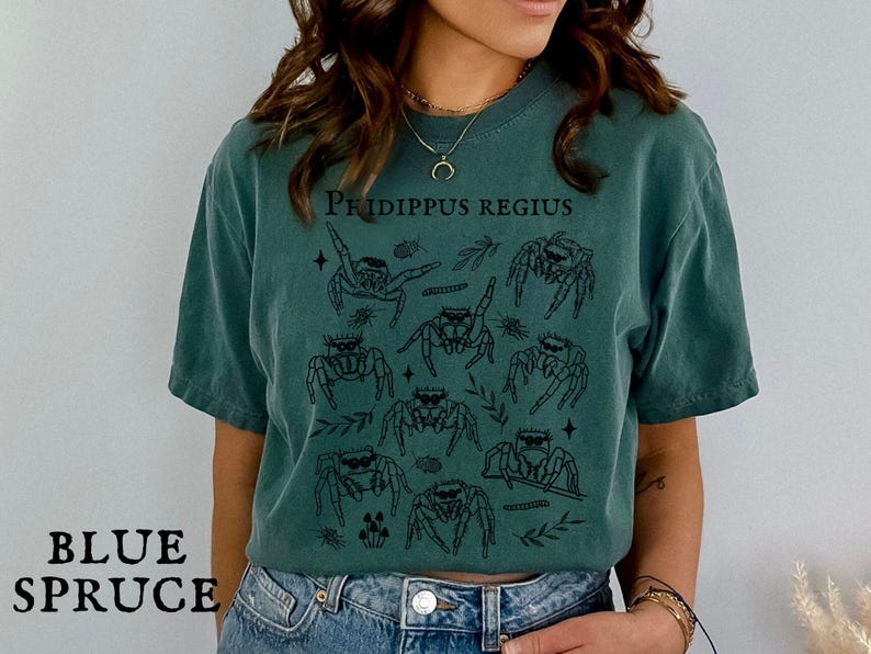 Pu&ograve; includere: T-shirt blu abete con un'illustrazione nera di ragni saltatori e la scritta "Phidippus Regius". Il design include elementi botanici. La maglietta ha una vestibilit&agrave; ampia.