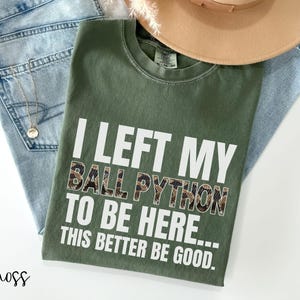May include: A moss green t-shirt with the text "I LEFT MY BALL PYTHON TO BE HERE... THIS BETTER BE GOOD." The shirt is folded on top of blue jeans, a gold necklace, and a tan hat.
