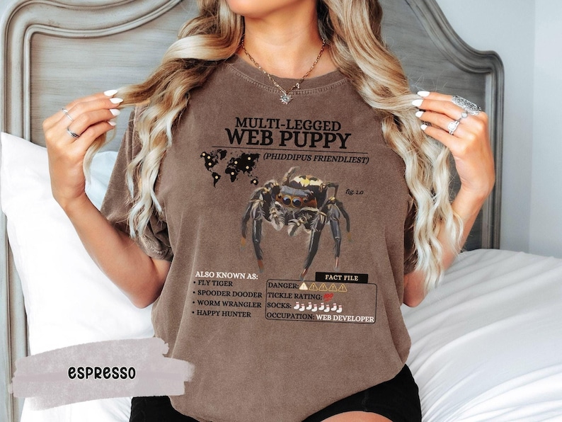 May include: Brown t-shirt with a graphic of a jumping spider and the text "MULTI-LEGGED WEB PUPPY (PHIDDIPUS FRIENDLIEST)". The text also includes "ALSO KNOWN AS: FLY TIGER, SPOODER DOODER, WORM WRANGLER, HAPPY HUNTER" and "FACT FILE: DANGER AAAAA, TICKLE RATING: SOCKS زززززززز, OCCUPATION: WEB DEVELOPER".