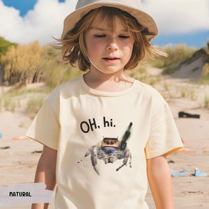 May include: A child wearing a light yellow t-shirt with a black and white jumping spider graphic and the text "OH. hi." The child is wearing a light brown hat and is standing on a sandy beach.