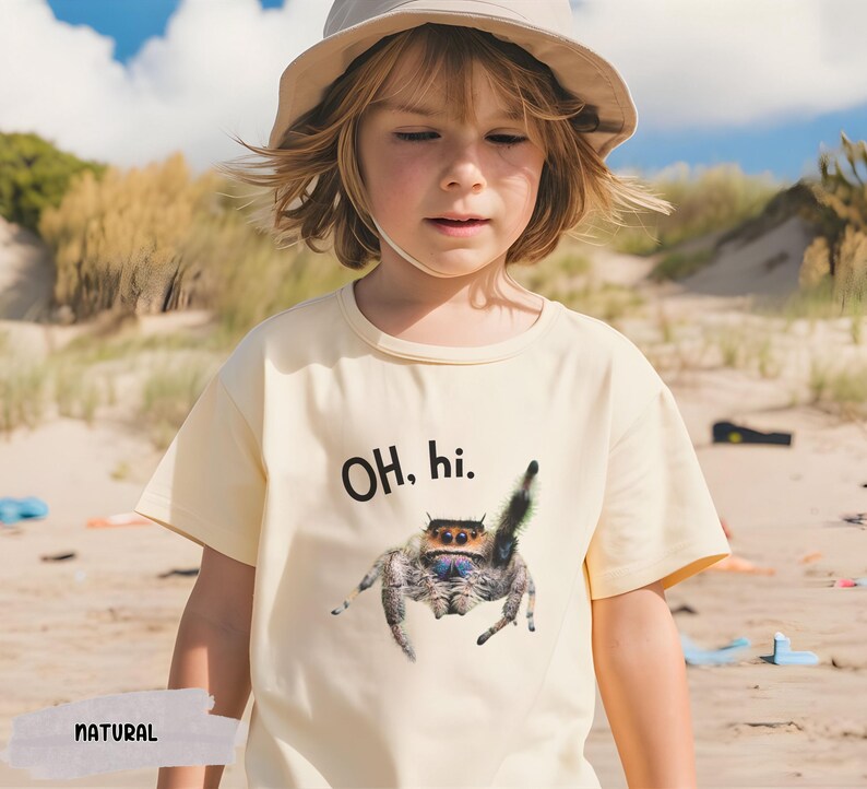 May include: A child wearing a light yellow t-shirt with a black and white jumping spider graphic and the text "OH. hi." The child is wearing a light brown hat and is standing on a sandy beach.
