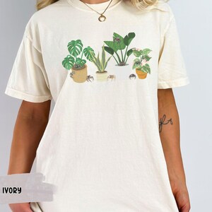 May include: A white t-shirt with a graphic of potted plants and spiders. The shirt is available in the color ivory.