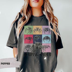 Op de afbeelding: Een donkergrijs T-shirt met een kleurrijke afbeelding van springende spinnen en de tekst "I'm My Jumping Spider Era".