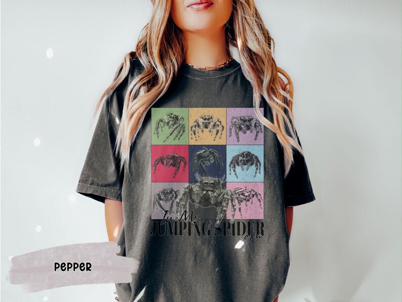 Op de afbeelding: Een donkergrijs T-shirt met een kleurrijke afbeelding van springende spinnen en de tekst "I'm My Jumping Spider Era".