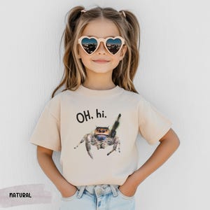 May include: A light beige t-shirt with short sleeves features a graphic of a jumping spider with the text "OH, hi." printed on the front. The shirt is being worn by a child with brown hair and heart-shaped sunglasses.
