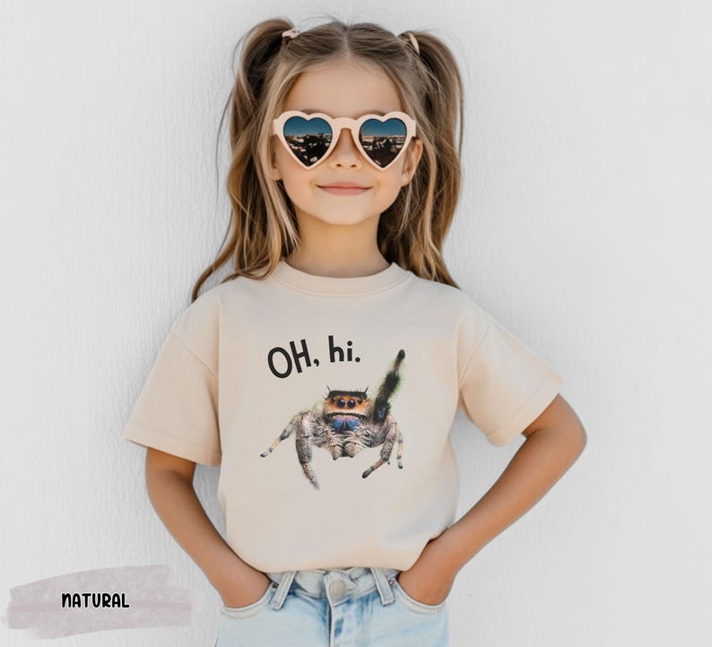 May include: A light beige t-shirt with short sleeves features a graphic of a jumping spider with the text "OH, hi." printed on the front. The shirt is being worn by a child with brown hair and heart-shaped sunglasses.