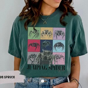 Op de afbeelding: Een donkergroen T-shirt met een kleurrijke afbeelding van springende spinnen en de tekst "I'm My Jumping Spider Era".