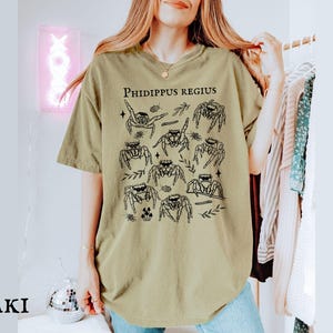 Pu&ograve; includere: T-shirt color cachi con un'illustrazione nera di diversi ragni saltatori ed elementi botanici. Il testo "Phidippus Regius" &egrave; stampato in alto. La maglietta ha una vestibilit&agrave; ampia.