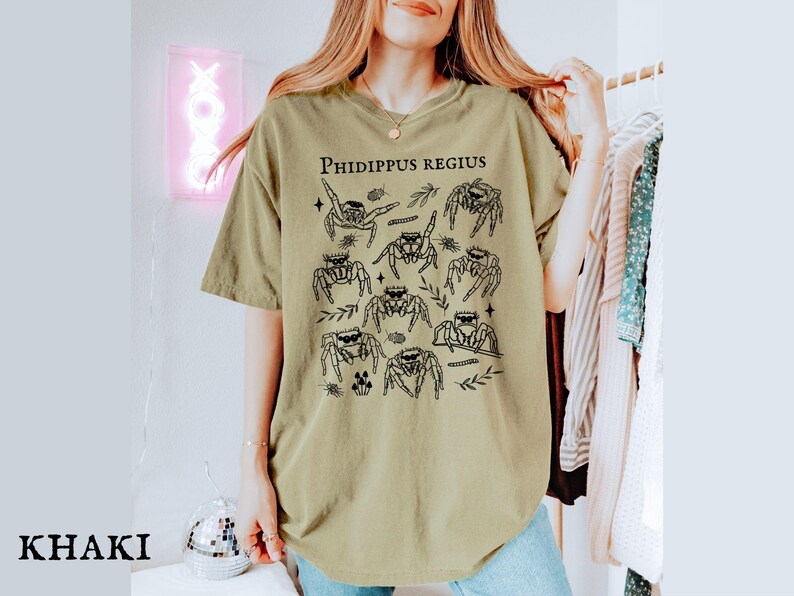 Pu&ograve; includere: T-shirt color cachi con un'illustrazione nera di diversi ragni saltatori ed elementi botanici. Il testo "Phidippus Regius" &egrave; stampato in alto. La maglietta ha una vestibilit&agrave; ampia.