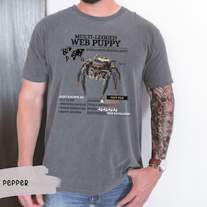 May include: A gray t-shirt with a black and white illustration of a jumping spider. The text "MULTI-LEGGED WEB PUPPY (PHIDDIPUS FRIENDLIEST)" is printed on the shirt. The shirt also includes a fact file about the spider, including its danger level, tickle rating, and occupation.
