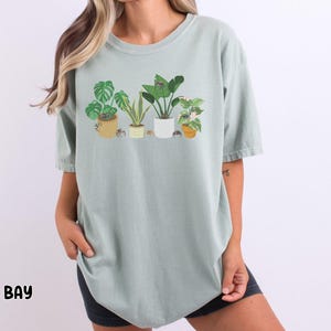 May include: A light green t-shirt with a graphic of potted plants. The plants are a variety of green and have brown pots. The t-shirt has a relaxed fit and a crew neck.