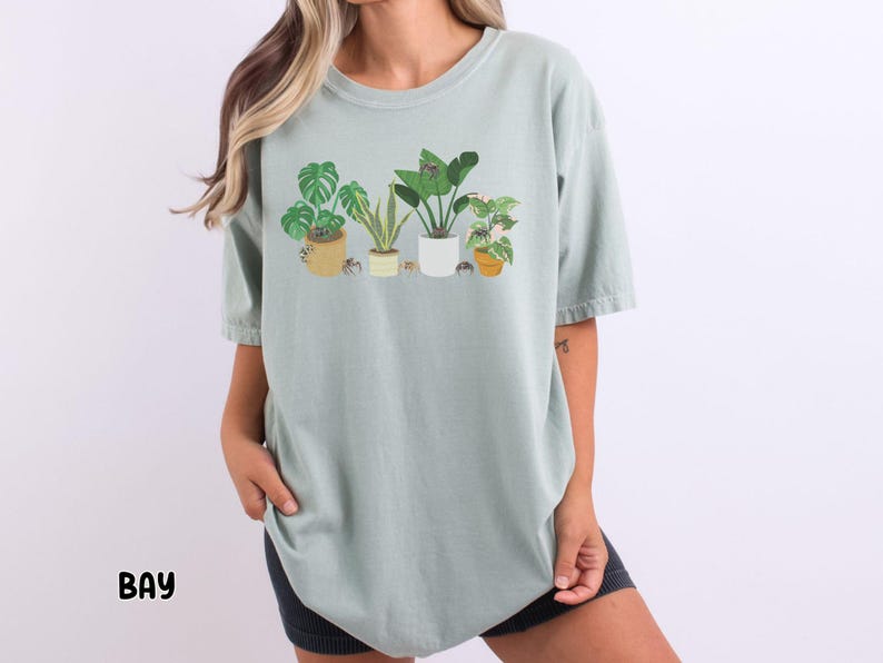 May include: A light green t-shirt with a graphic of potted plants. The plants are a variety of green and have brown pots. The t-shirt has a relaxed fit and a crew neck.