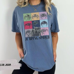 Op de afbeelding: Een blauw jeans T-shirt met een afbeelding van springende spinnen in kleurrijke vierkanten. De tekst "In My Jumping Spider Era" is op het shirt gedrukt.