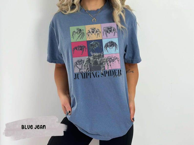 Op de afbeelding: Een blauw jeans T-shirt met een afbeelding van springende spinnen in kleurrijke vierkanten. De tekst "In My Jumping Spider Era" is op het shirt gedrukt.