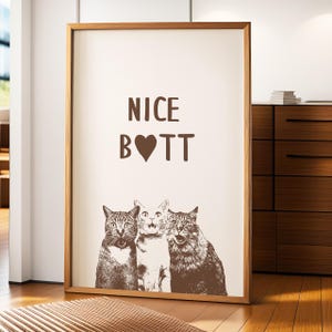 May include: A framed print with the text "NICE B♡TT" in brown lettering on a white background. Three cats are pictured below the text, looking up at the viewer.