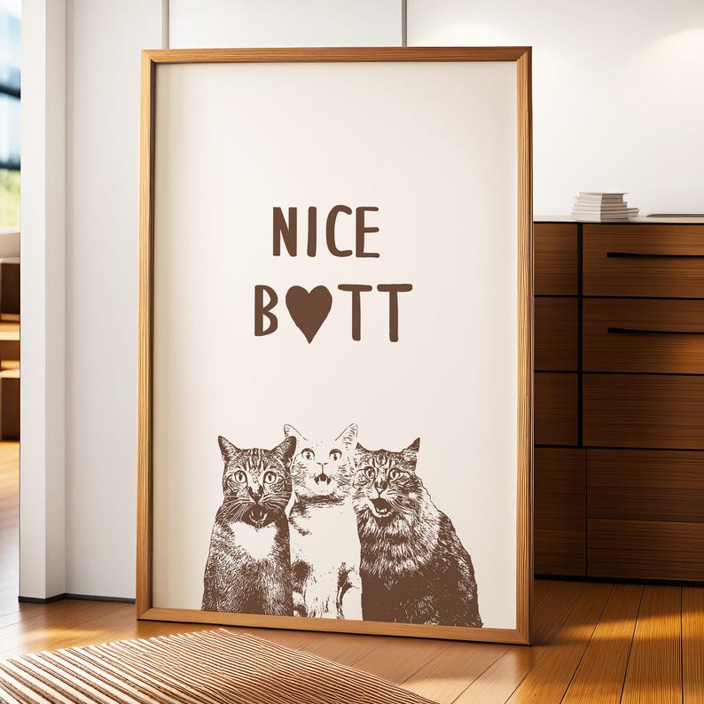May include: A framed print with the text "NICE B♡TT" in brown lettering on a white background. Three cats are pictured below the text, looking up at the viewer.